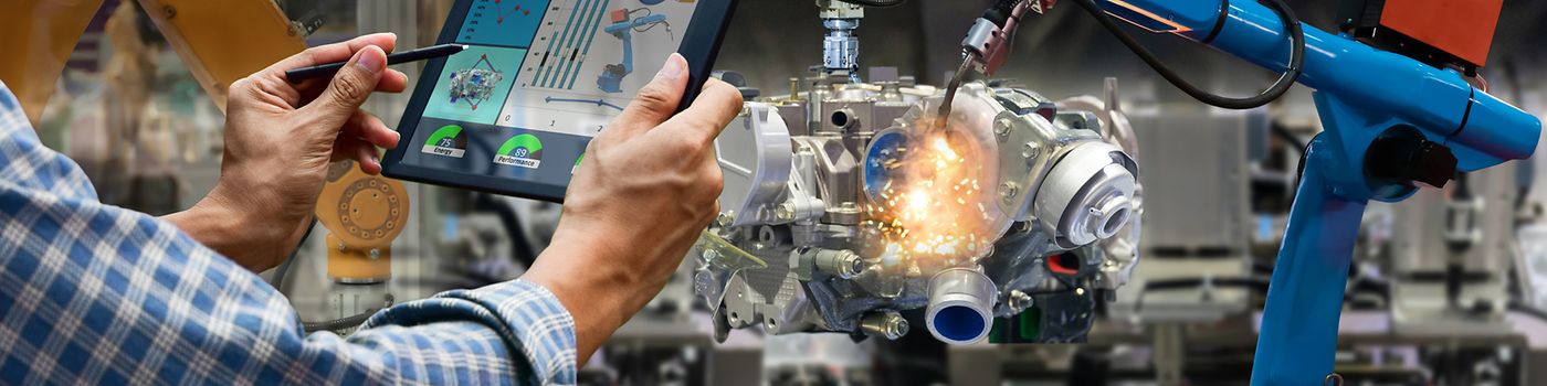 Engineer on factory floor with tablet and robot arms in background